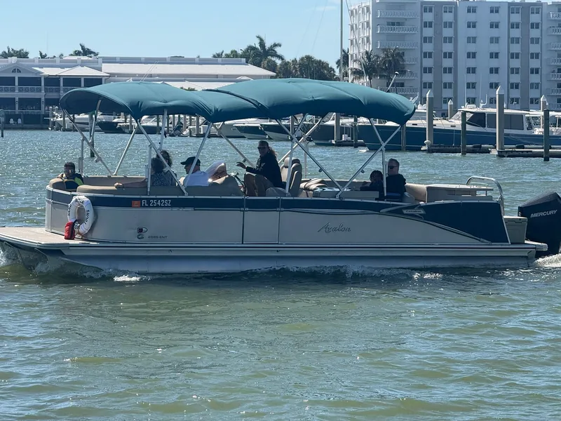 The teal-canopied Sand Dollar Avalon pontoon underway on Naples Bay, passing a marina of docked boats with low condo buildings behind
