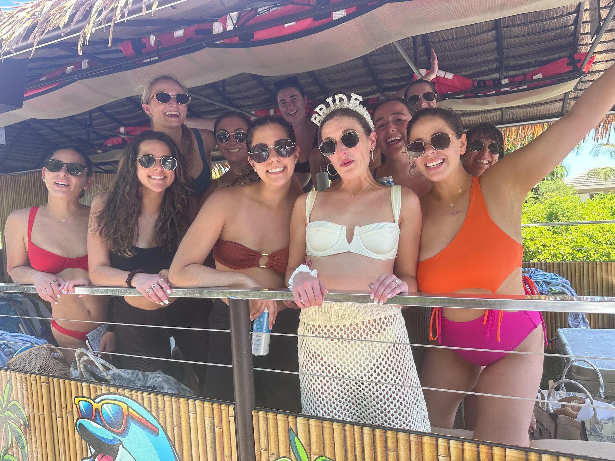 A bachelorette group in colorful swimsuits leaning on the tiki boat rail, the bride wearing a pearl BRIDE crown, Marco Island, Florida