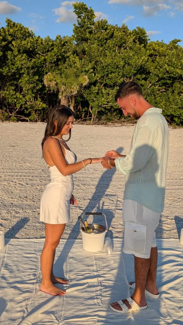 A man slipping a ring onto his partner's finger on a picnic blanket set with a chilled bottle on the beach