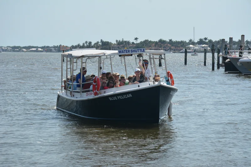 The Blue Pelican water shuttle coming bow-on across Naples Bay loaded with guests, a palm-lined shoreline behind it