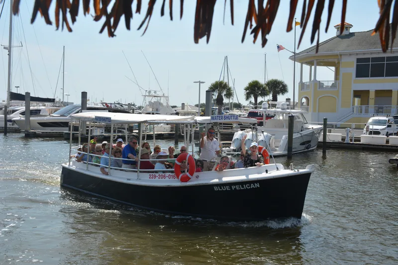 Guests waving from our Blue Pelican water shuttle at the Naples Bay marina, sailboat masts and a yellow marina building behind them