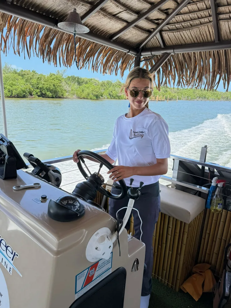 A Sand Dollar crew member in a white Sand Dollar Shelling shirt steering the tiki boat under its thatched roof, mangroves and the boat's wake behind her