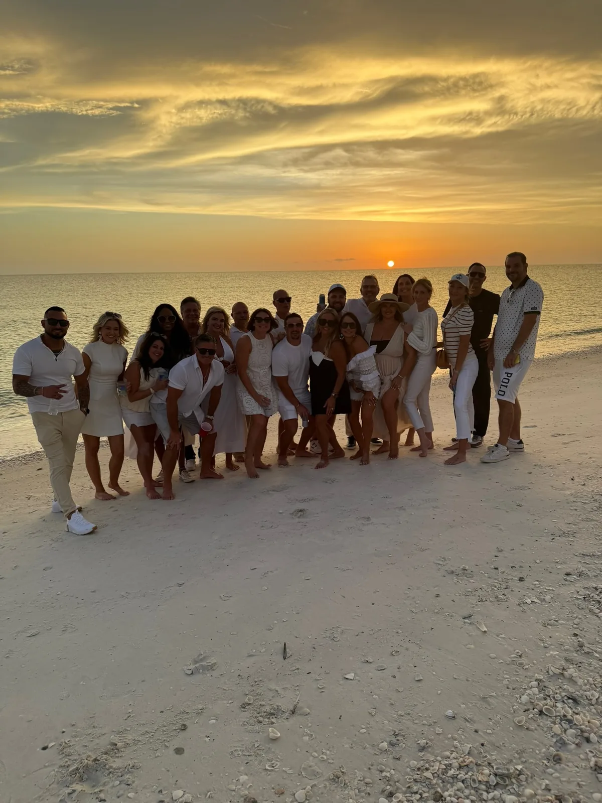 A large group dressed in white posing together on a shell-covered beach under a golden sunset