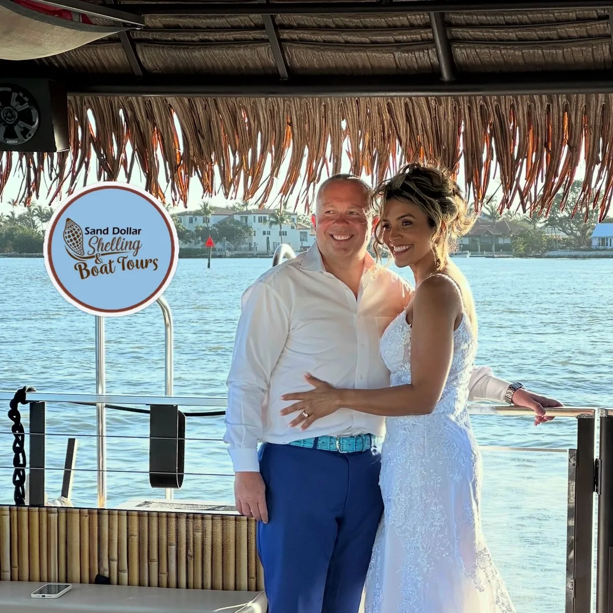 A bride and groom under the tiki boat's thatched roof beside the round Sand Dollar Shelling and Boat Tours sign