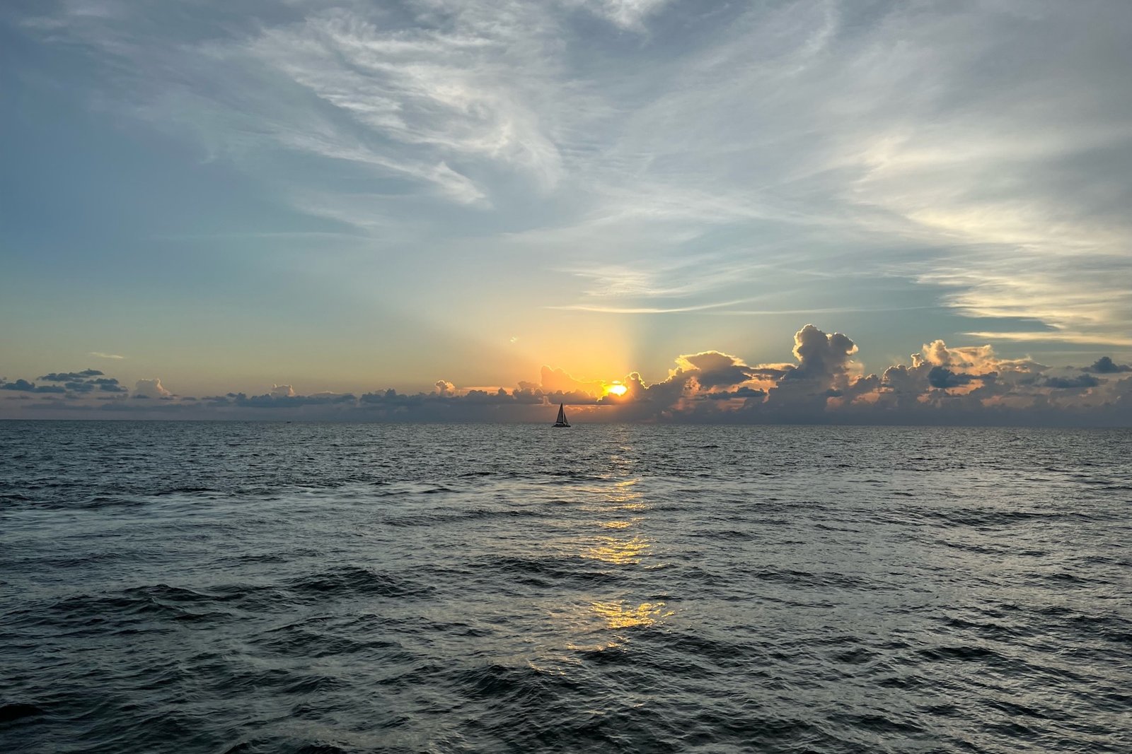 The sun dropping toward the Gulf of Mexico horizon with a distant sailboat and gold light on the water off Naples, Florida