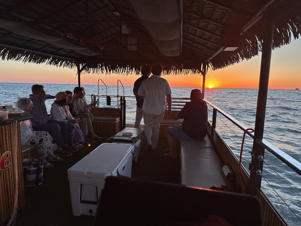 Guests under the thatched roof of a Sand Dollar tiki boat watching the sun drop into the Gulf near Naples, Florida